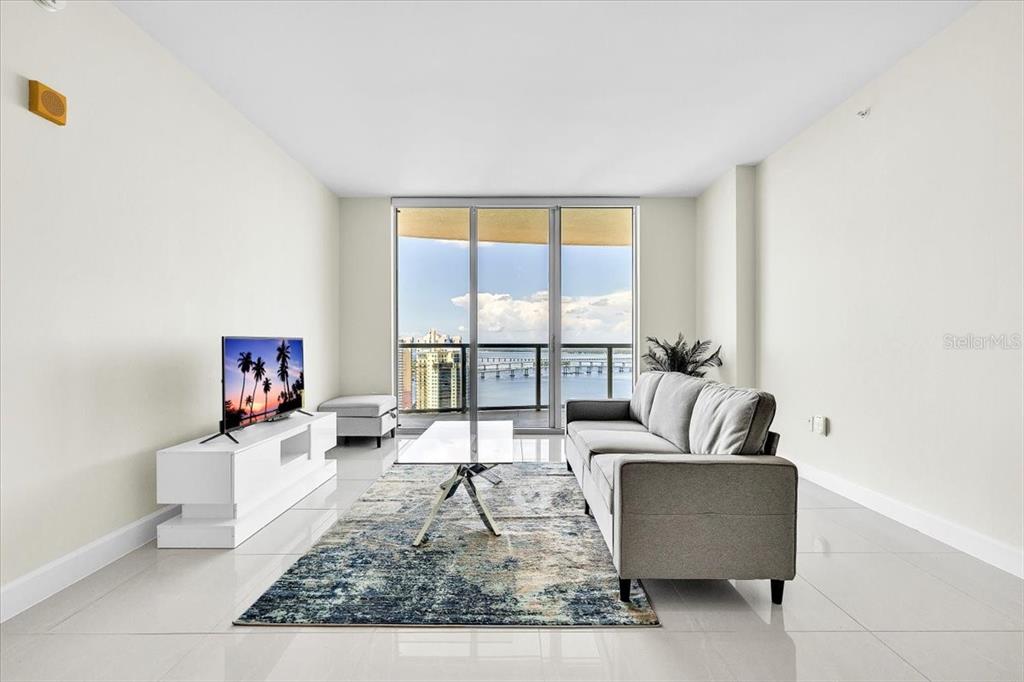 A modern living room features a gray sofa, white media console, and a balcony with water views — 3000 OASIS GRAND BOULEVARD UNIT 2802, FORT MYERS, FL