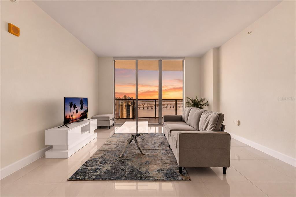 A living room with a grey sofa, glass coffee table, and a view of the water — 3000 OASIS GRAND BOULEVARD UNIT 2802, FORT MYERS, FL