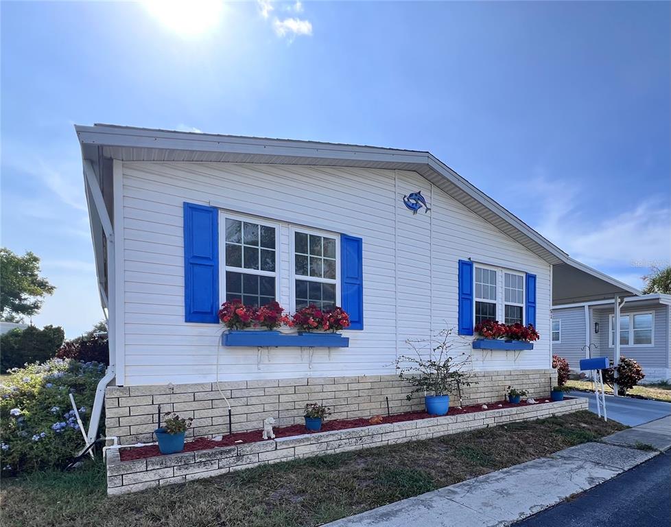 A white manufactured home features blue shutters, window flower boxes, and a brick-patterned foundation — 51 PINDO PALM STREET E, LARGO, FL