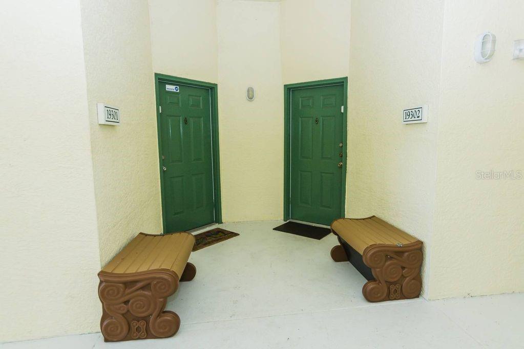 Two green doors with benches sit on a concrete porch between beige stucco walls — 8923 LEGACY COURT UNIT 302, KISSIMMEE, FL
