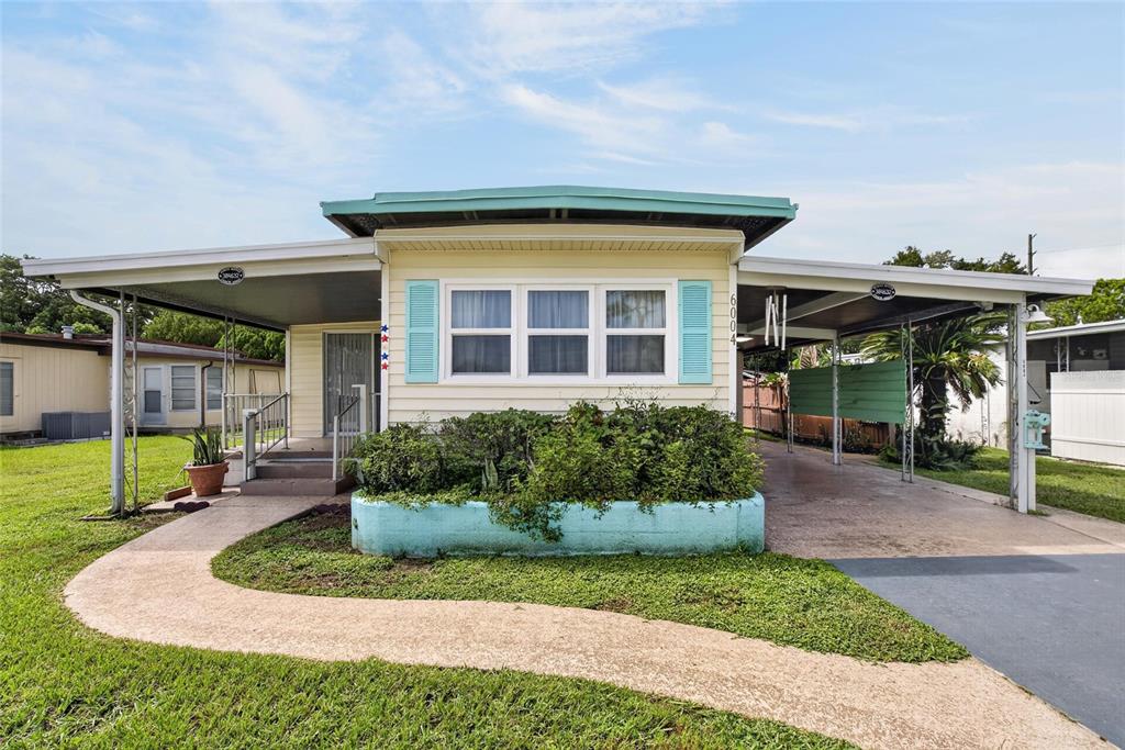 A single-story mobile home with a turquoise planter, teal shutters, and a covered carport at sunset — 6004 ALPINE DRIVE, ZEPHYRHILLS, FL