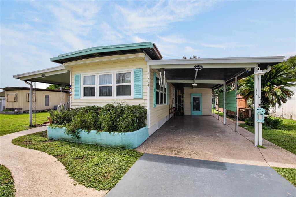A single-story manufactured home features a light yellow exterior, blue shutters, and a covered carport — 6004 ALPINE DRIVE, ZEPHYRHILLS, FL