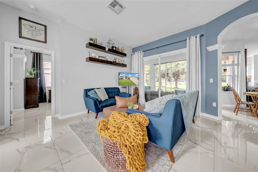 A living room features blue sofas, floating shelves, and tile flooring with a view outside — 4455 GEVALIA DRIVE, BROOKSVILLE, FL