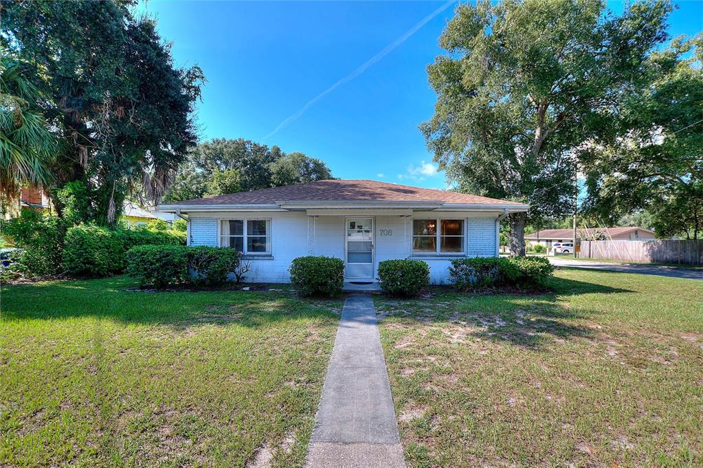 A single-story white brick house sits on a grassy lot surrounded by large trees — 708 AVENUE E NE, WINTER HAVEN, FL