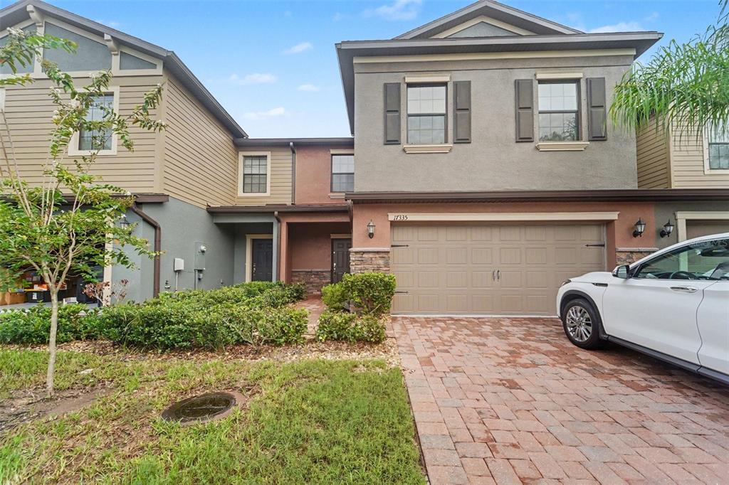 A two-story townhouse with a tan garage, brick paver driveway, and a small front yard — 17335 OLD TOBACCO ROAD, LUTZ, FL