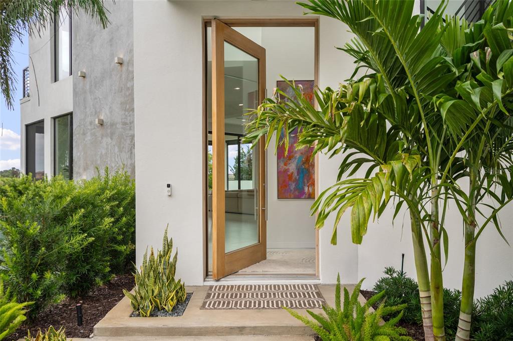 A modern home entrance features a wooden pivot door, a patterned doormat, and tropical landscaping — 9061 RON DEN LANE, WINDERMERE, FL