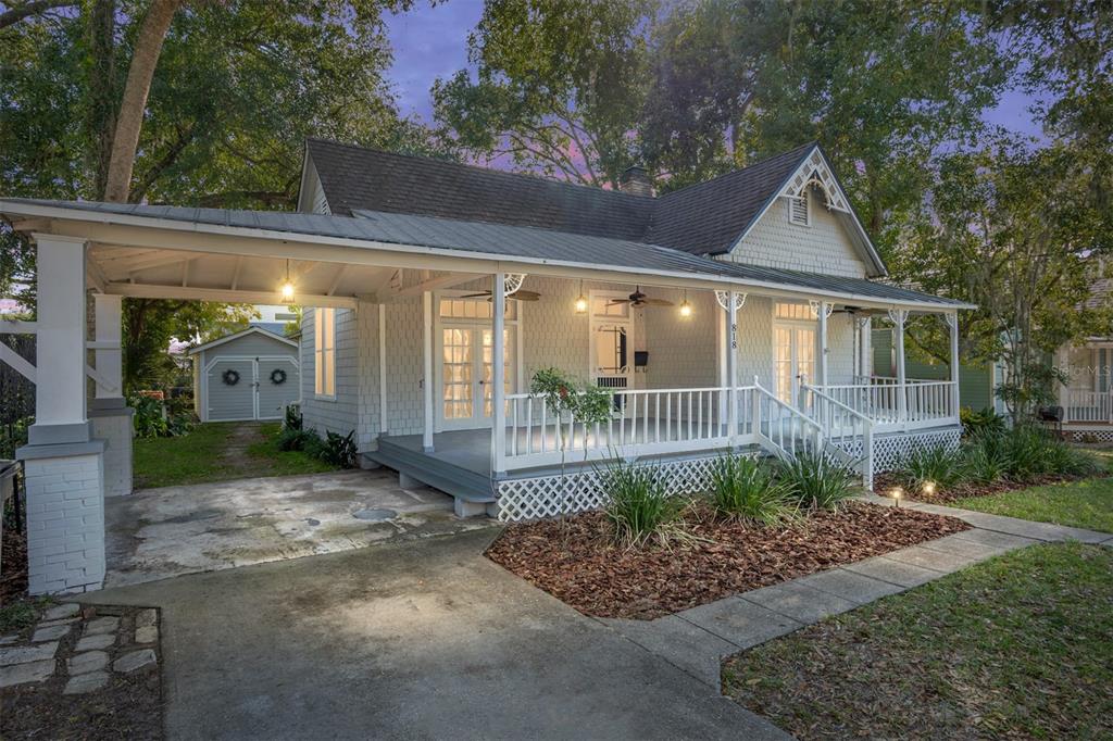 A light-colored cottage with a wrap-around porch, metal roof, and attached carport at dusk — 818 SE 3RD STREET, OCALA, FL