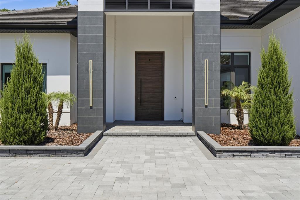 A modern home entrance features a dark wood door, gray stone pillars, and two tall evergreens — 15800 PENDIO DRIVE, BELLA COLLINA, FL