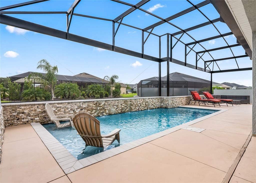 A screened-in patio features a rectangular swimming pool with two submerged chairs and stone wall — 3146 JACQUELINE WAY, THE VILLAGES, FL