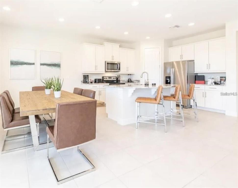 A bright, open-concept kitchen and dining area featuring white cabinetry, stainless appliances, and brown chairs — 7437 MARKER AVENUE, KISSIMMEE, FL