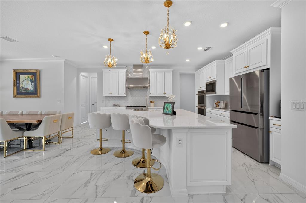 A modern white kitchen features a marble-topped island, gold bar stools, and three crystal pendant lights — 33751 LANDSMAN LOOP, WESLEY CHAPEL, FL