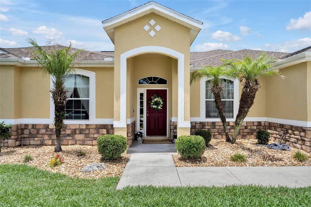 A tan single-story house features a red front door, stone siding, and two palm trees — 4946 NE 124TH ROAD, OXFORD, FL