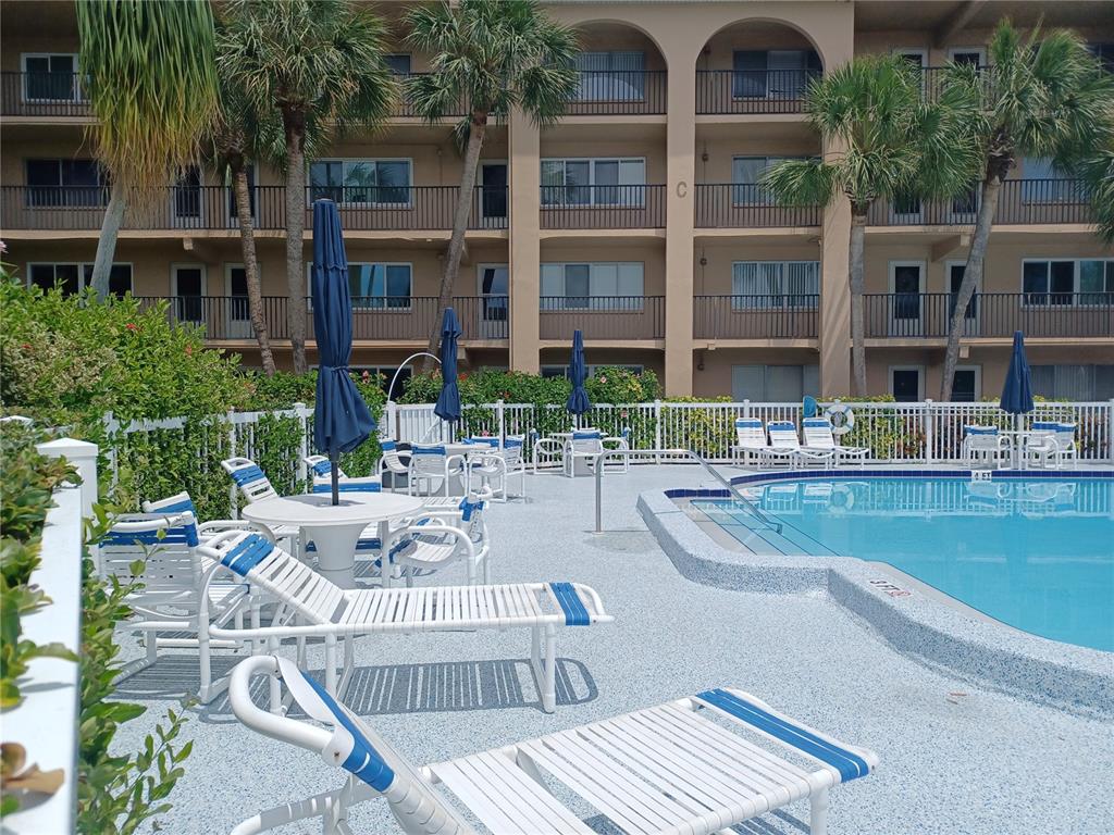 A swimming pool area with lounge chairs, tables, umbrellas, and a multi-story apartment building — 2525 W BAY DRIVE UNIT A12, BELLEAIR BLUFFS, FL