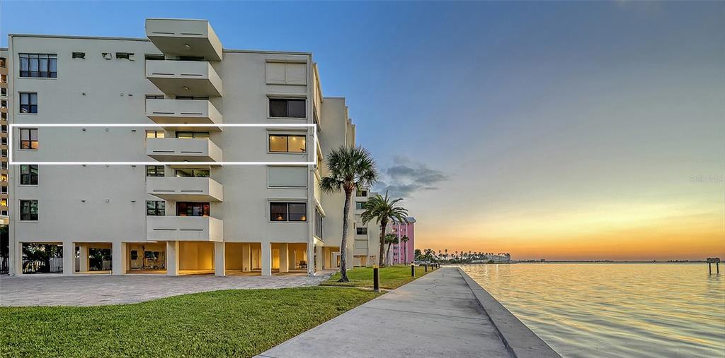 A multi-story apartment building stands beside a concrete walkway along the water at sunset — 101 SUNSET DRIVE UNIT 103, SARASOTA, FL