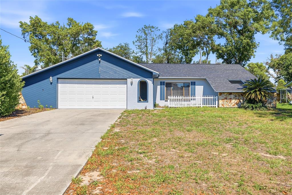 A single-story house with a blue garage, stone accents, and a white picket fence — 3276 MORVEN DRIVE, SPRING HILL, FL