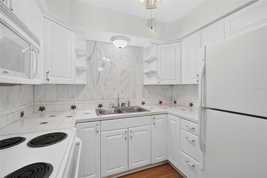 A white kitchen features tile countertops, a double sink, and a marble-look tile backsplash — 138 36TH AVENUE N UNIT 208 W, ST PETERSBURG, FL