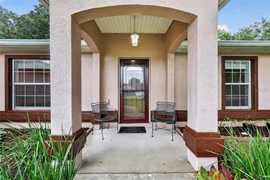 A covered front porch features two metal chairs, a storm door, and beige stucco walls — 8825 SE 155TH PLACE, SUMMERFIELD, FL