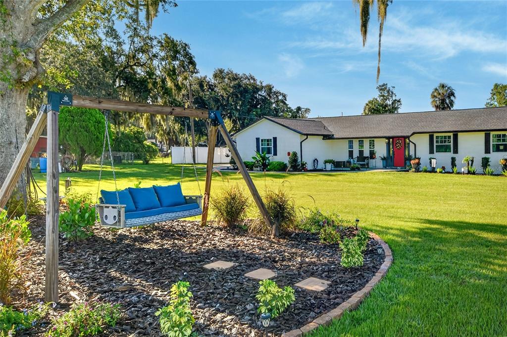A wooden swing with blue cushions sits in a mulched garden before a white house — 4220 HOWELL LANE, MULBERRY, FL