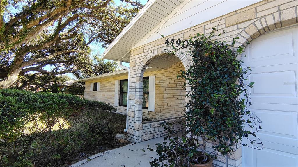 A stone-faced house exterior features an arched entryway, a white garage door, and overgrown landscaping — 4790 MICHAEL LANE, PONCE INLET, FL