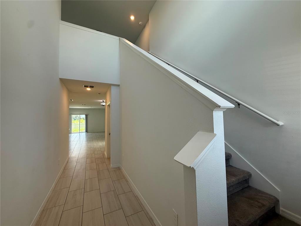 A hallway with tile flooring leads toward a windowed room beside a carpeted staircase — 35190 GRAVELLY DRIVE, ZEPHYRHILLS, FL