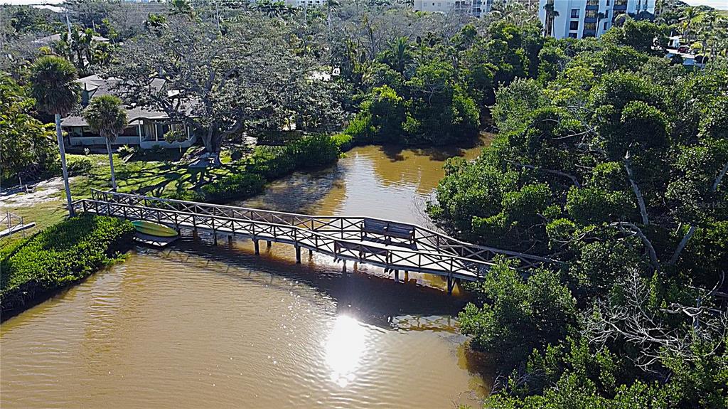 A wooden footbridge crosses a narrow, murky waterway surrounded by lush green trees and vegetation — 8534 HERON LAGOON CIRCLE, SARASOTA, FL