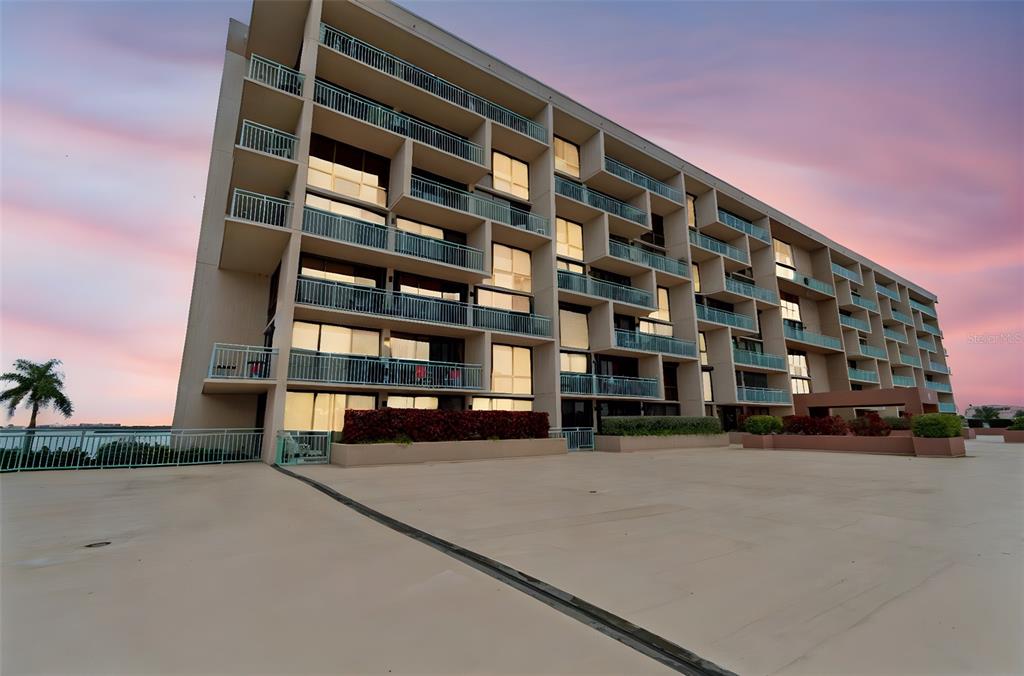 A multi-story apartment building with balconies stands above a large, paved outdoor deck at sunset — 1 KEY CAPRI UNIT 101E, TREASURE ISLAND, FL