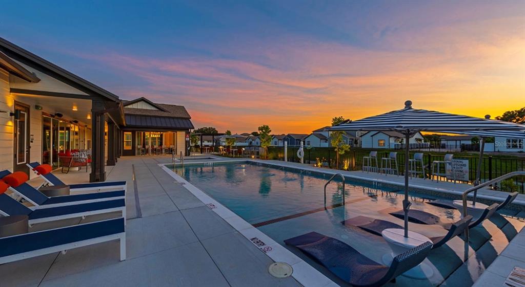 A swimming pool with lounge chairs and an umbrella sits beside a modern clubhouse building — 4980 RED MAPLE LANE UNIT C1, WILDWOOD, FL