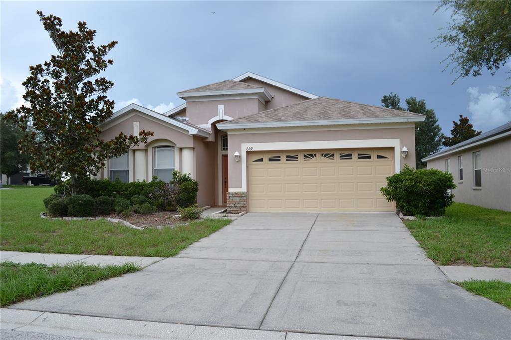 A single-story house exterior features a tan stucco facade, a beige garage door, and an arched entryway — 659 WINTHROP DRIVE, SPRING HILL, FL