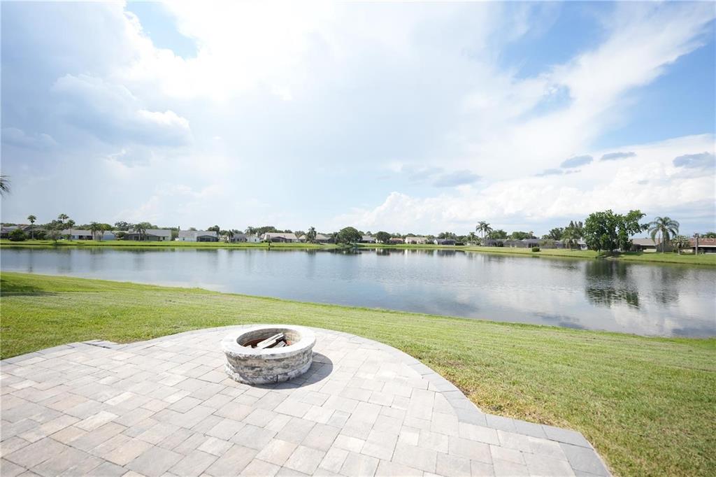 A stone fire pit sits on a paved patio overlooking a lake and grassy yard — 14712 EAGLES CROSSING DRIVE, ORLANDO, FL