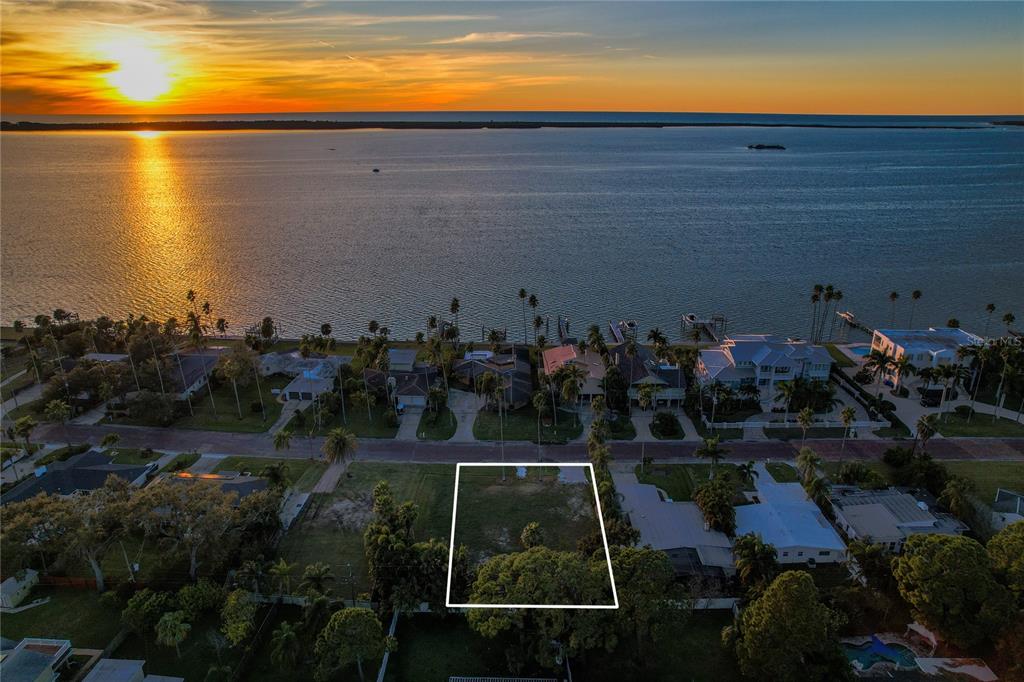 An aerial view shows a vacant residential lot outlined in white near a waterfront sunset — 1645 SANTA BARBARA DRIVE, DUNEDIN, FL