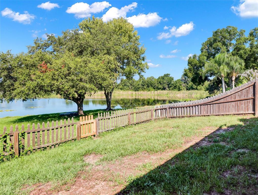 A grassy backyard features a wooden picket fence, a small gate, and a pond view — 22817 SAINT THOMAS CIRCLE, LUTZ, FL