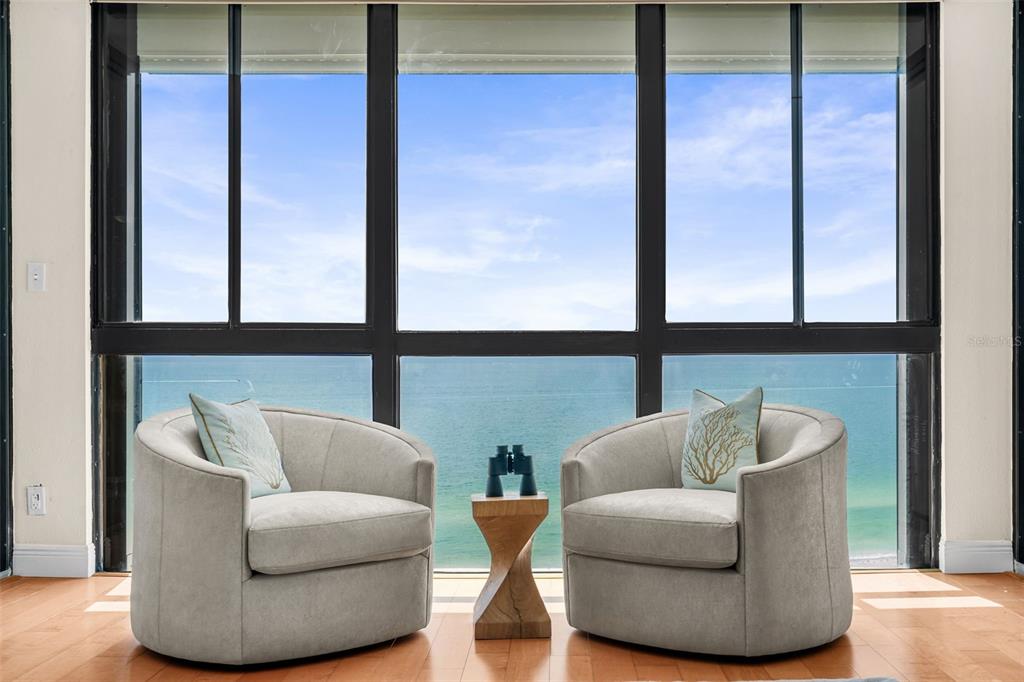 Two gray armchairs and a small wooden table sit before a large window overlooking ocean — 3820 GULF BOULEVARD UNIT 1205, ST PETE BEACH, FL