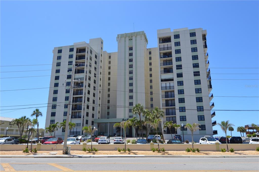 A tall, multi-story apartment building with balconies stands behind a parking lot and palm trees — 3820 GULF BOULEVARD UNIT 1205, ST PETE BEACH, FL