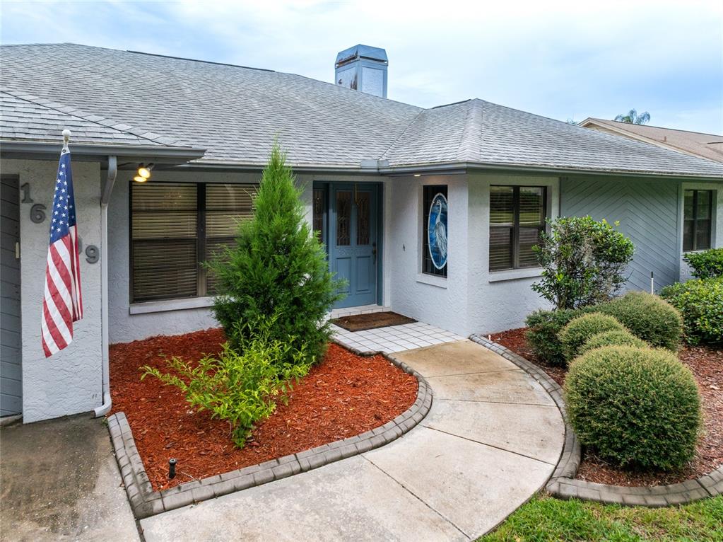 A single-story house exterior features a blue front door, a flag, and a landscaped walkway — 16509 E COURSE DRIVE, TAMPA, FL