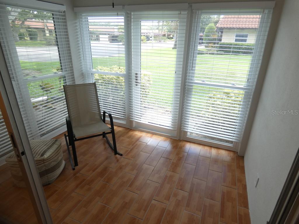 A sunroom features wood-look tile flooring, a single chair, and large windows with white blinds — 1701 PINEHURST ROAD UNIT 9G, DUNEDIN, FL