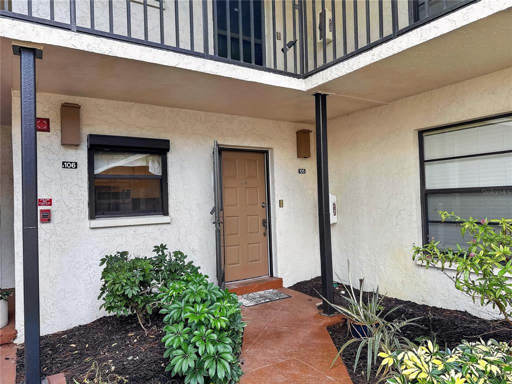 An exterior view of a ground-level apartment entrance with a brown door and landscaping — 1701 COURTYARD WAY UNIT 105, NAPLES, FL