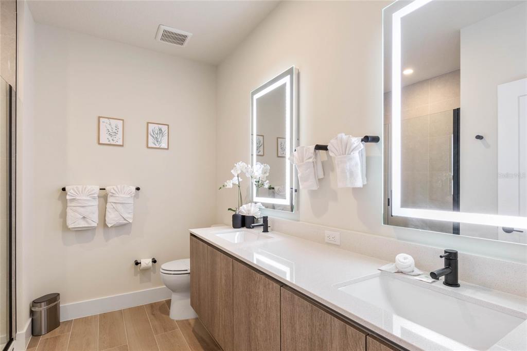 A modern bathroom features a double vanity with light wood cabinets and two illuminated mirrors — 17244 BLESSING DRIVE, CLERMONT, FL