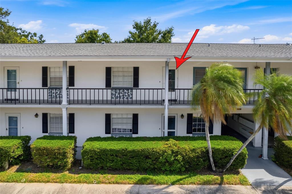 A two-story apartment building exterior features a second-floor balcony with a red arrow pointing toward a door — 100 GLENNES LANE UNIT 204, DUNEDIN, FL