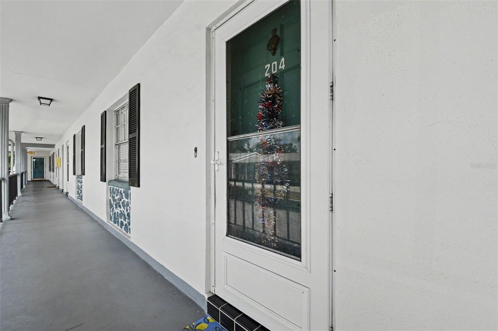 A long outdoor walkway features white walls, black shutters, and a door marked 204 — 100 GLENNES LANE UNIT 204, DUNEDIN, FL