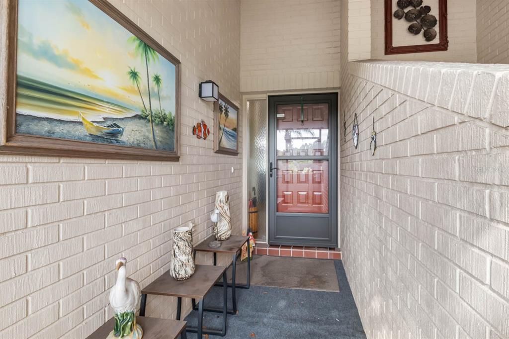 A covered entryway features a brick wall, a storm door, and decorative wall art — 2519 LONIGAN PLACE, SUN CITY CENTER, FL