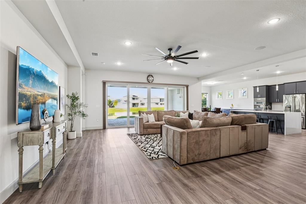 An open-concept living room features wood-look flooring, a large sectional sofa, and a kitchen — 8743 SANDERS TREE LOOP, WESLEY CHAPEL, FL