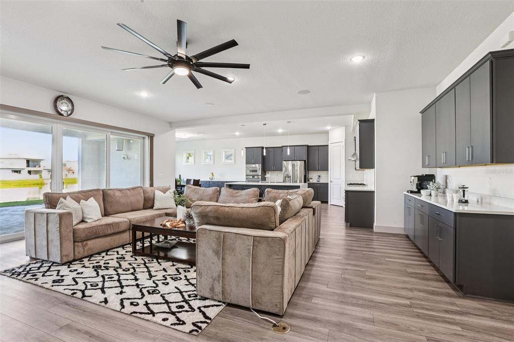 An open-concept living room and kitchen featuring gray cabinetry, wood-look flooring, and a large sectional — 8743 SANDERS TREE LOOP, WESLEY CHAPEL, FL