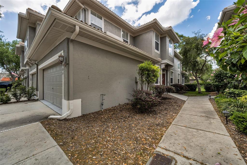 A gray two-story townhouse exterior features a garage, a concrete walkway, and landscaped garden beds — 3304 COVERED BRIDGE DRIVE E, DUNEDIN, FL