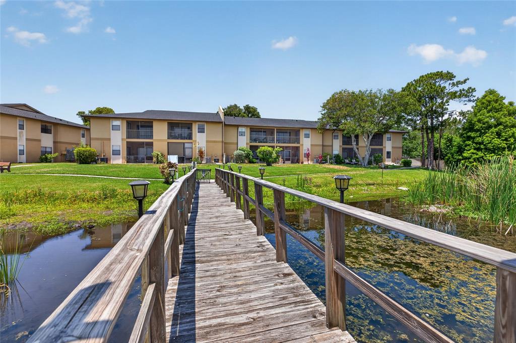A wooden footbridge leads toward a two-story apartment building overlooking a small pond — 10125 SAILWINDS BOULEVARD N UNIT 104, LARGO, FL