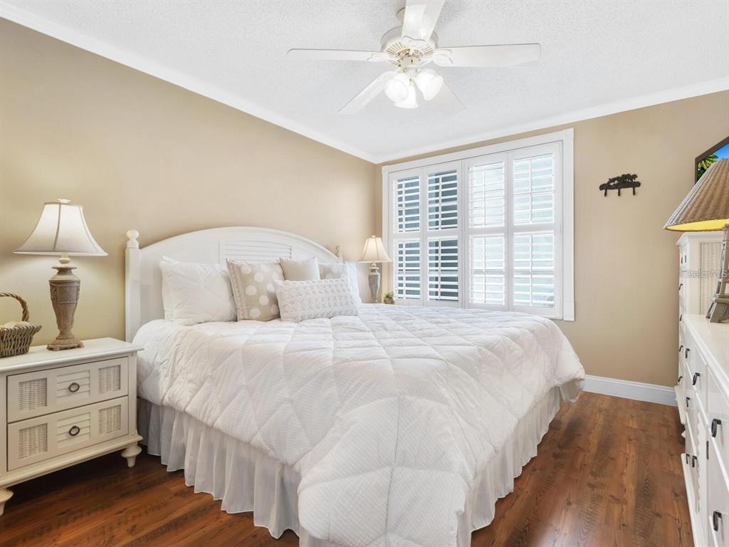 A bedroom features a white bed, wooden floors, plantation shutters, and a ceiling fan — 600 SUTTON PLACE UNIT 201, LONGBOAT KEY, FL