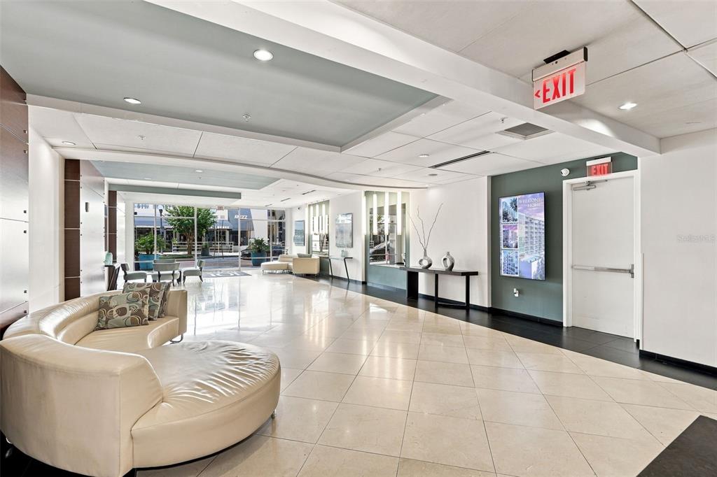 A modern building lobby features a curved white sofa, tiled floors, and an exit sign — 101 S EOLA DRIVE UNIT 905, ORLANDO, FL