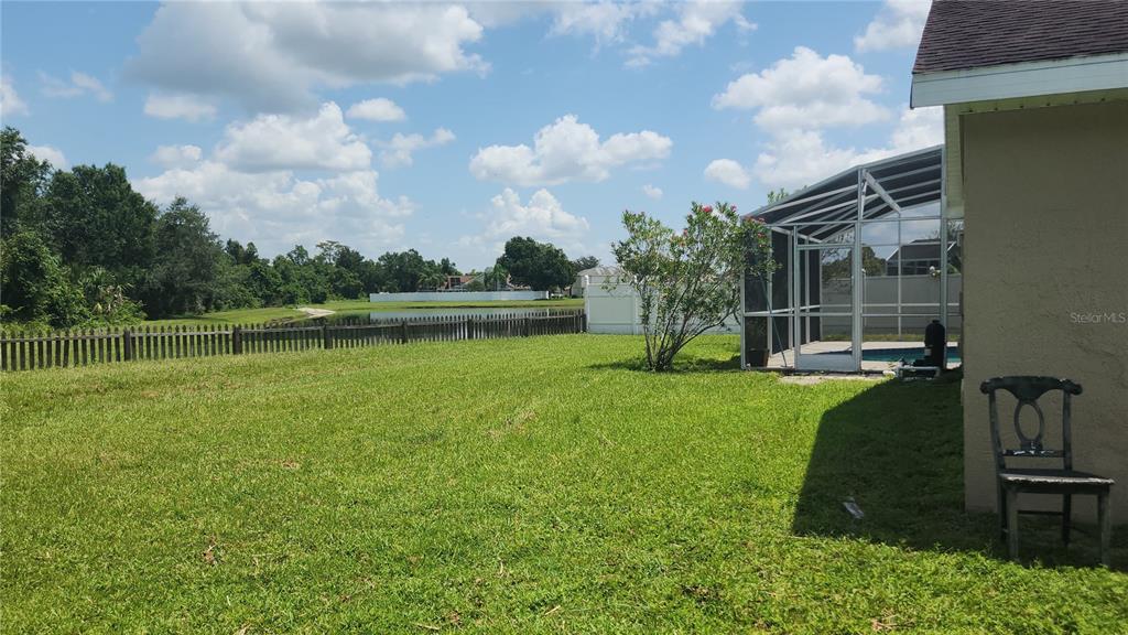 A grassy backyard features a screened-in pool enclosure, a wooden fence, and a distant pond — 3119 FAIRFIELD DRIVE, KISSIMMEE, FL