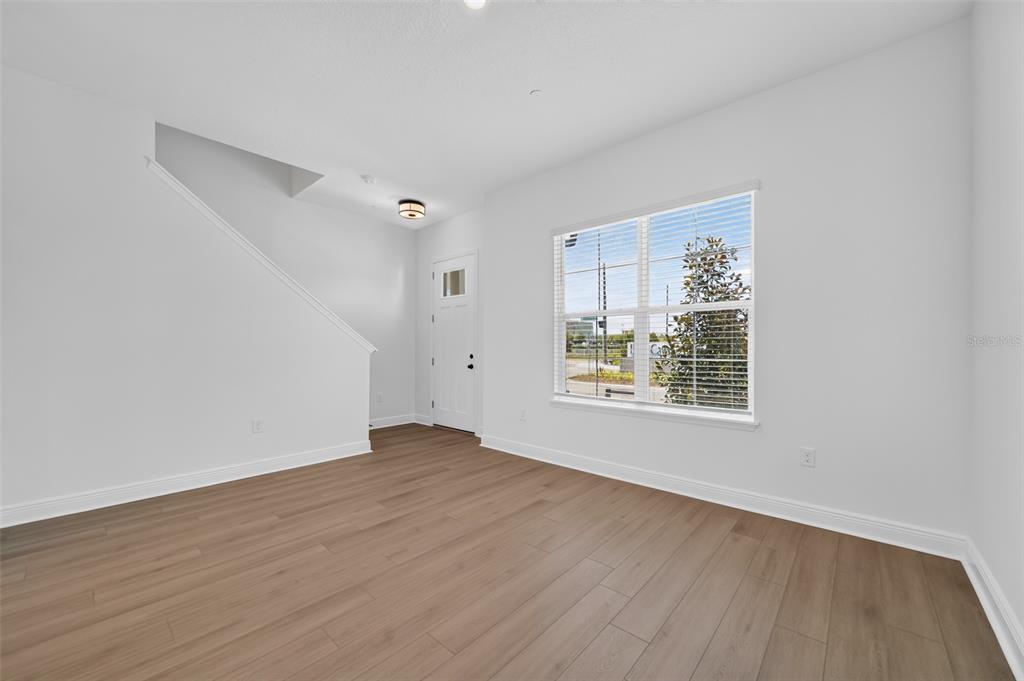 An empty room features light wood-look flooring, white walls, a staircase, and a large window — 13321 LUSH LIME ALLEY, WINDERMERE, FL