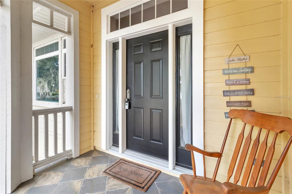 A yellow-sided front porch features a black door, a welcome mat, and a wooden rocking chair — 1215 LAKESHORE DRIVE, EUSTIS, FL