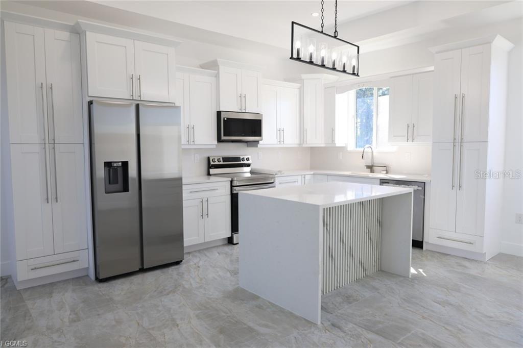 A modern white kitchen features stainless steel appliances, a marble-accented island, and light tile flooring — 114 PRESTON STREET, LEHIGH ACRES, FL